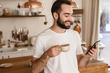Portrait of laughing brunette man 20s drinking coffee and using smartphone at home