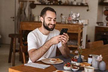 Portrait of beautiful brunette man 20s using smartphone while having breakfast at home