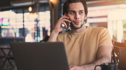 Young man talking mobile phone and using laptop computer in cafe