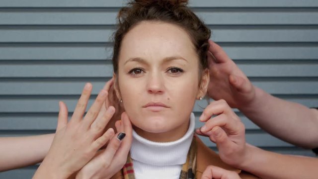 Portrait of afraid young business woman who is touched by plenty of hands against the background of a striped wall. Sad brunette girl close up among the palms of hands. Social concept