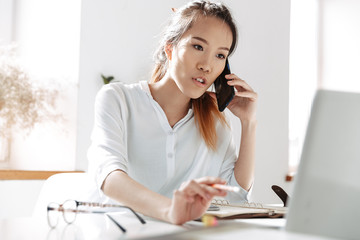 Obraz premium Concentrated asian business woman talking by smartphone and using laptop