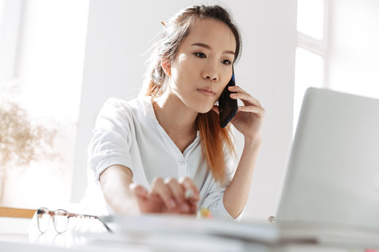 Serious Asian Business Woman Talking By Smartphone And Using Laptop