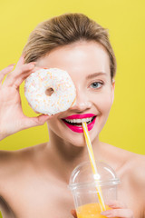 Obraz na płótnie Canvas cheerful woman covering eye with tasty doughnut while holding straw near mouth isolated on yellow