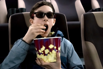 The teenager in the cinema in 3D glasses with popcorn in their hands. The guy opened his mouth and watches the movie in surprise.