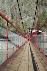 suspension bridge in the mountains with a wooden floor on the background of the house with a girl walking on it with a backpack