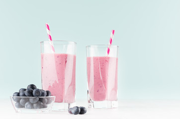 Two detox milk violet drinks with striped straws, ripe blueberry in bowl on soft pastel blue background and white wood table, copy space.