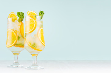 Summer fresh cold cocktails with oranges, green mint, ice, straw on soft light pastel green background and white wood table.