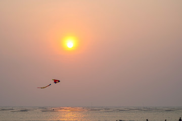 View from the beach with a yellow glowing sun setting in the horizon with a flying kite.