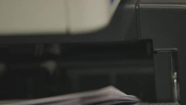 Handheld, interior, extreme close up shot of a printer printing out paper. The tray lowers and raises up and down.
