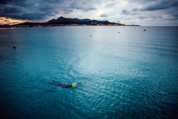 Female swimmer swim in open water. Dark sky, and mountains in background.