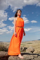 Young woman in orange long dress outdoor. Rocks and heaven background.