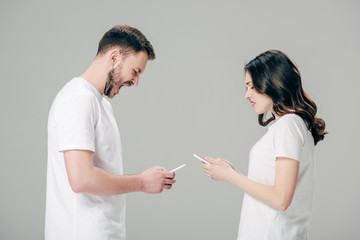 Fototapeta premium handsome man using smartphone and yelling while standing near displeased girl with smartphone isolated on grey