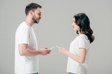 Fototapeta premium young man and woman in white t-shirts looking at each other while holding smartphones isolated on grey