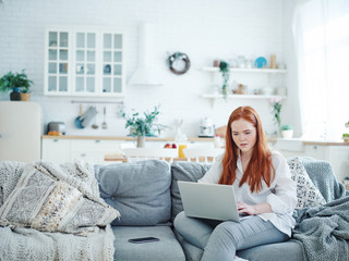 Beautiful young red-haired woman sitting on sofa at home and working on laptop looking confused
