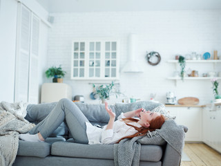 Side view of young woman with red hair lying on sofa at home and talking on cell phone with friend