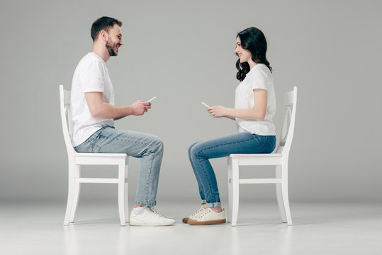 side view of smiling man and woman in white t-shirts and blue jeans sitting on chairs with smartphones and looking at each other on grey background