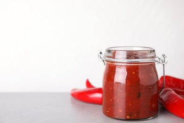 Glass jar of hot chili sauce with peppers on table against white background. Space for text