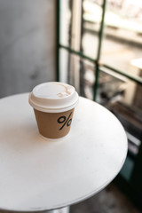 Close up composition of paper hot coffee cup on white metal table setting near glass window in natural light scene / coffee shop concept / nice composition background / background for advertising