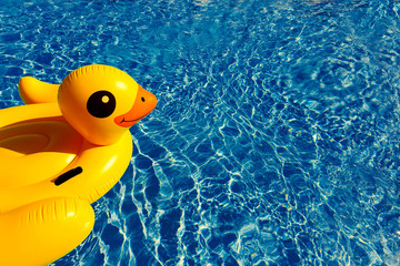 Bright yellow inflatable swimming duck in ripple blue water in swimming pool of resort hotel with sun reflection. Summer holiday concept, background with copy space.