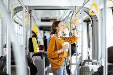 Young woman passenger enjoying trip at the public transport, standing with coffee in the modern tram - obrazy, fototapety, plakaty