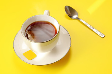 Morning sun shines at freshly brewed steaming cup of tea on yellow board, bag still in, silver spoon next to it
