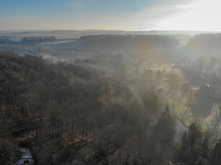 Obraz premium Aerial view of foggy and cold winter morning with blue sky facing the sun over the forest and farmland in Belgium, Walloon Brabant