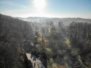 Fototapeta premium Aerial view of foggy and cold winter morning with blue sky facing the sun over the forest and farmland in Belgium, Walloon Brabant