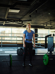 Fototapeta na wymiar Strong young woman wearing weightlifting belt standing by barbell with plates preparing to do exercise in gym