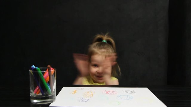 Little girl shows her hands and claps her hands while sitting on a dark gray sofa at the table during a drawing class.