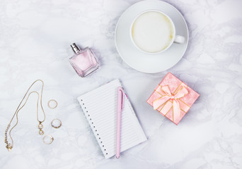 Women's accessories on a white marble table. Notepad, pen and glass of coffee on the table and other female cosmetic accessories. Beauty and fashion concept. Top view, flat lay, copy space