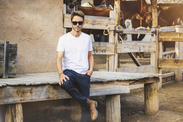 Young handsome bearded man is staying on a western city background surrounded by wooden fence. Guy is wearing a white empty t-shirt without logo. Horizontal mock-up style.