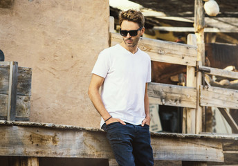 Young handsome bearded man is staying on a western city background surrounded by wooden fence. Guy is wearing a white empty t-shirt without logo. Horizontal mock-up style.