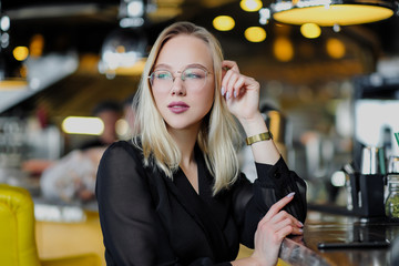 A young beautiful blond woman with glasses sits at the bar in a yellow interior. Drinks alcoholic cocktail with a straw and green mint.