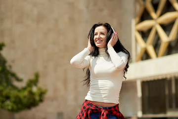 Street hip hop dancer on sunny day. Young happy sportive woman in urban background listening music in white wireless headphones and dancing. Hair fluttering in the wind