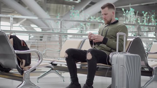 Young man is untangling wires of headphones, sitting in terminal station