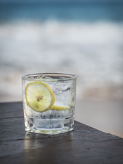 Glass of gin with tonic, lemon and ice cubes on the wooden table. Sea on the background.