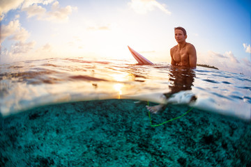 Surfer sits on the surfboard and looks at the camera at sunset. Splitted view with underwater view