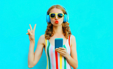 Portrait cool girl blowing red lips sending sweet air kiss holding phone listening to music in wireless headphones on colorful blue background
