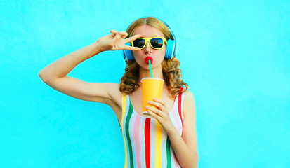 Portrait cool girl drinking fruit juice listening to music in wireless headphones on colorful blue background - obrazy, fototapety, plakaty