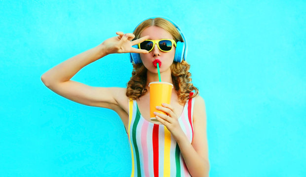 Portrait cool girl drinking fruit juice listening to music in wireless headphones on colorful blue background