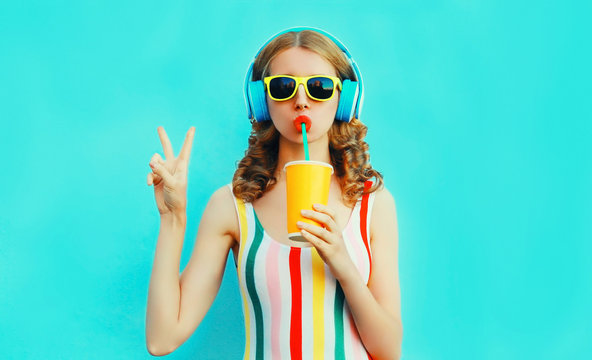 Portrait cool girl drinking fruit juice listening to music in wireless headphones on colorful blue background