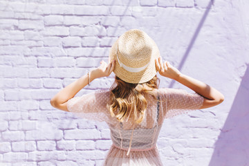 Close-up portrait from back of slim romantic girl in light dress looking at old wall. Outdoor photo of blonde curly lady in straw hat with ribbon posing on brick background.