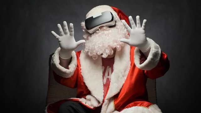 Santa Claus is having fun with glasses of virtual reality sitting in a chair on a black background.