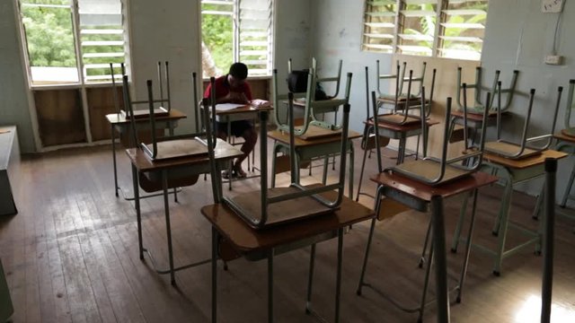Steady, medium wide shot of a student doing work at a desk. The other chairs are turned upside down on top of desks.