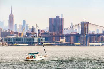 View of midtown Manhattan skyline, Brooklyn Bridge and sailboat that is cruising on East River during sunny summer day in New York City, USA