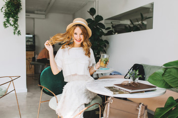 Cheerful girl in trendy vintage hat playing with blonde hair and laughing relaxing in cafe. Excited fair-haired lady in white outfit drinking cocktail and having fun while chilling in cozy restaurant.