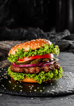 Beef burger with tomatoes, red onions, cucumber and lettuce on black slate over dark background. Unhealthy food.