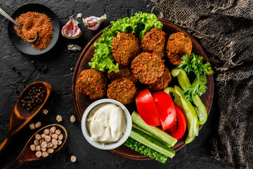 Roasted chickpeas falafel patties with garlic yogurt sauce, served with lettuce and fresh vegetables in a plate over dark stone background. Healthy vegan food, clean eating, dieting, top view