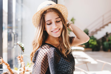 Lovely fair-haired girl with big happy smile posing in light room enjoying good memories. Indoor close-up portrait of charming young woman in hat and wristwatch laughing with eyes closed.