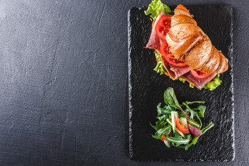 Croissant sandwich with prosciutto, tomatoes, cheese parmesan and greens on black shale board over black stone background. Healthy food concept, top view.